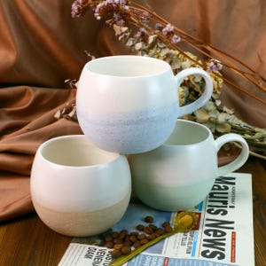 Puede incluir: Tres tazas de cerámica con un esmalte moteado. Las tazas están apiladas una encima de la otra. La taza de arriba es azul claro, la taza del medio es verde claro y la taza de abajo es beige.