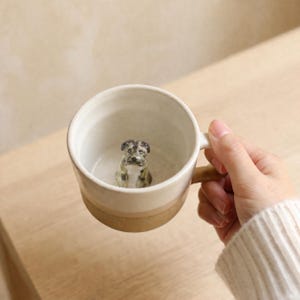 May include: A cream ceramic mug with a brown base and handle. Inside the mug is a small, detailed figurine of a dog. The dog is primarily grey and white, with a focused expression. The mug is held by a person.