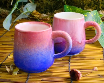 Op de afbeelding: Twee keramische mokken met een kleurovergang van rood naar blauw. De mokken hebben afgeronde handvatten en witte binnenkanten. Ze staan op een bamboemat, met groen op de achtergrond. Een unieke toevoeging voor elke keuken.