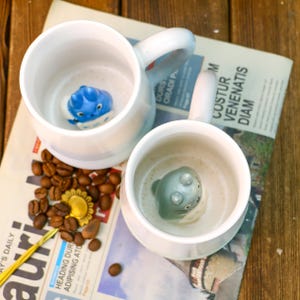 May include: Two white ceramic mugs with blue and gray 3D animal figures inside. The mugs have a light blue and white striped design. The mugs are sitting on a newspaper with coffee beans scattered around them.