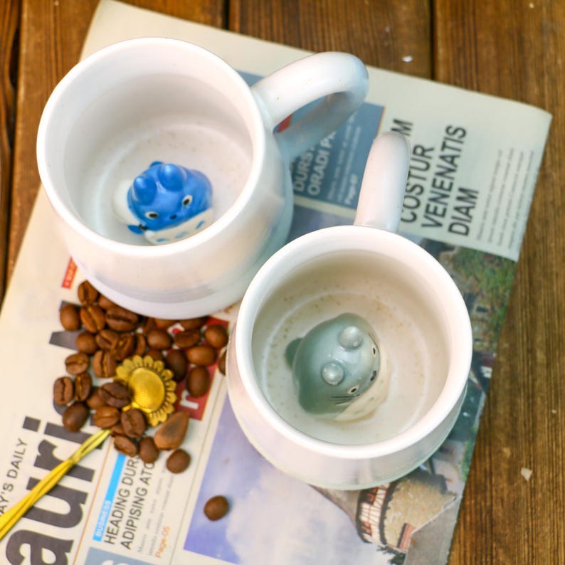 May include: Two white ceramic mugs with blue and gray 3D animal figures inside. The mugs have a light blue and white striped design. The mugs are sitting on a newspaper with coffee beans scattered around them.