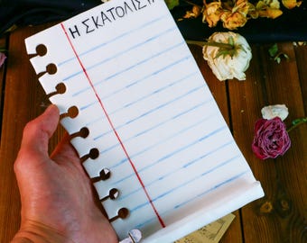 May include: A white ceramic notepad with blue lines and a red margin, held by a hand. The notepad has the Greek text "Η ΣΚΑΤΟΛΙΣΤΑ" at the top. The notepad has a rolled-up bottom edge and a series of holes along the left side.