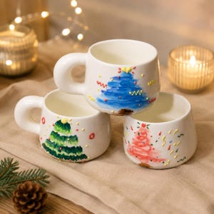 May include: Three white ceramic mugs with Christmas tree designs. One mug features a blue tree, another a green tree, and the third a red tree. Each mug has a rounded handle and colorful accents.