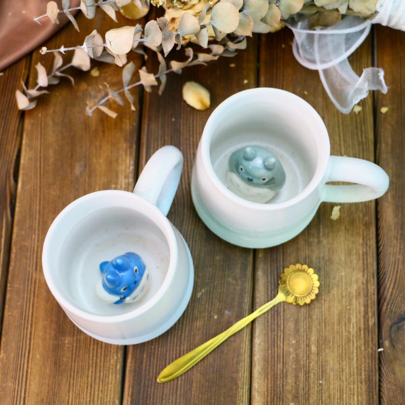 May include: Two white ceramic mugs with blue and gray character figurines inside. The mugs have a light blue ombre glaze. A gold spoon with a flower design is on the wooden table.