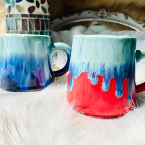 May include: Two ceramic mugs with a drip glaze design. One mug is blue and green, the other is red and green.