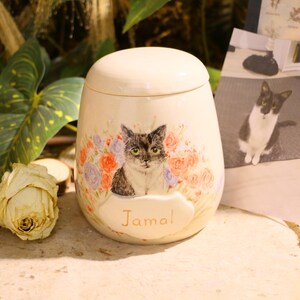 Puede incluir: Una urna de cer&aacute;mica de color crema con tapa redondeada, que presenta una ilustraci&oacute;n pintada a mano de un gato rodeado de flores naranjas y moradas. Debajo del gato, el nombre Jamal est&aacute; escrito en letras doradas sobre una placa en relieve.