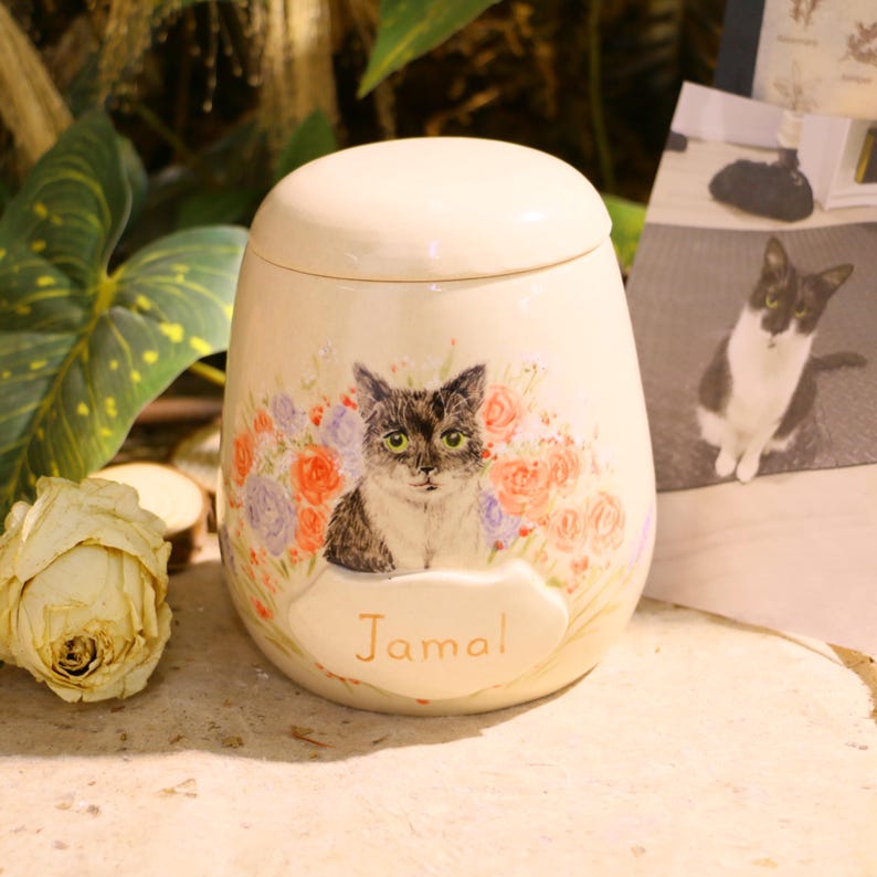 Puede incluir: Una urna de cer&aacute;mica de color crema con tapa redondeada, que presenta una ilustraci&oacute;n pintada a mano de un gato rodeado de flores naranjas y moradas. Debajo del gato, el nombre Jamal est&aacute; escrito en letras doradas sobre una placa en relieve.