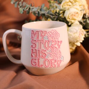 May include: White ceramic mug with a pink and blue floral design and the text "My Story His Glory".