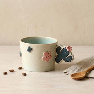 Puede incluir: Una taza de cerámica blanca con un asa negra y el texto "But First, Coffee" en negro y negrita. La taza está sobre una superficie blanca. La taza probablemente tiene 325 ml.
