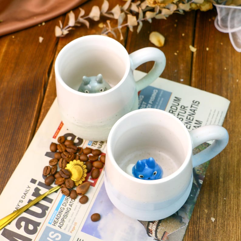 May include: Two ceramic mugs with blue and white glaze. Each mug has a small animal figurine inside. The mugs are sitting on a newspaper with coffee beans scattered around them.