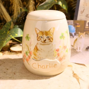 Puede incluir: Una urna de cer&aacute;mica color crema con tapa, que presenta un gato atigrado naranja pintado rodeado de peque&ntilde;as flores rosas y hojas de tr&eacute;bol verdes. El nombre Charlie est&aacute; escrito en letras marrones sobre una placa ovalada en relieve.