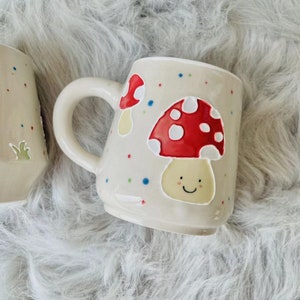 May include: Two ceramic mugs with a white background and colourful polka dots. One mug features a red and white mushroom with a smiling face. The other mug features a red and white mushroom.