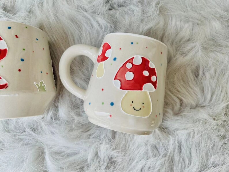 May include: Two ceramic mugs with a white background and colourful polka dots. One mug features a red and white mushroom with a smiling face. The other mug features a red and white mushroom.