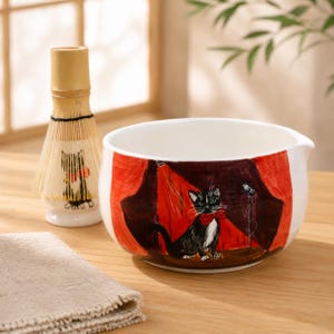 Könnte beinhalten: Eine weiße Matcha-Schale aus Keramik mit einem bemalten schwarzen Kater mit roter Fliege auf einer Bühne mit roten Vorhängen und einem Mikrofon, dazu ein Matcha-Besen aus Bambus mit einer passenden schwarzen Katzenillustration.