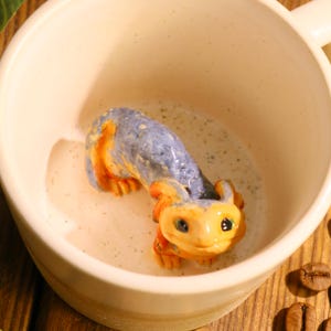 Puede incluir: Una taza de cerámica color crema con una criatura colorida esculpida en su interior. La criatura es azul, naranja y amarilla, y parece estar nadando en un líquido lechoso. Granos de café son visibles en la superficie de madera.