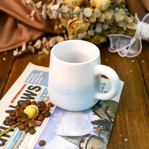May include: A white ceramic mug with a light blue glaze on the bottom half. The mug has a rounded shape and a handle.