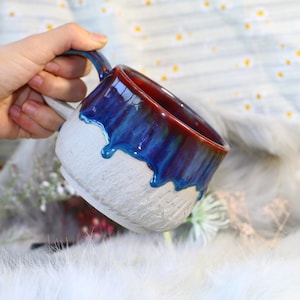 May include: A white ceramic mug with a blue and red glaze. The glaze has a drip effect, creating a unique and artistic look.