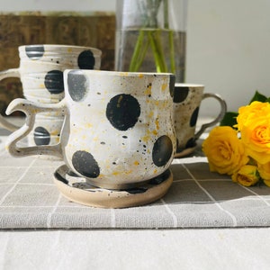 Könnte beinhalten: Handgefertigte Keramikbecher mit schwarzen Punkten und gelben Spritzern. Jeder Becher hat einen einzigartigen Griff und steht auf einer passenden Untertasse. Ideal für Kaffee oder Tee.
