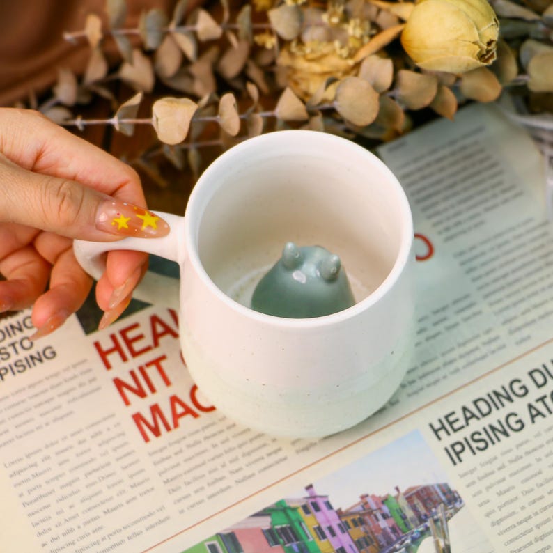 May include: White ceramic mug with a light green ceramic animal figurine inside. The mug has a handle and a speckled glaze.