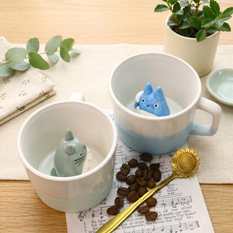May include: Two ceramic mugs with cartoon character figures inside. One mug is light blue and white, the other is white and light green. A gold spoon with a sunflower design and coffee beans are on a sheet of music.