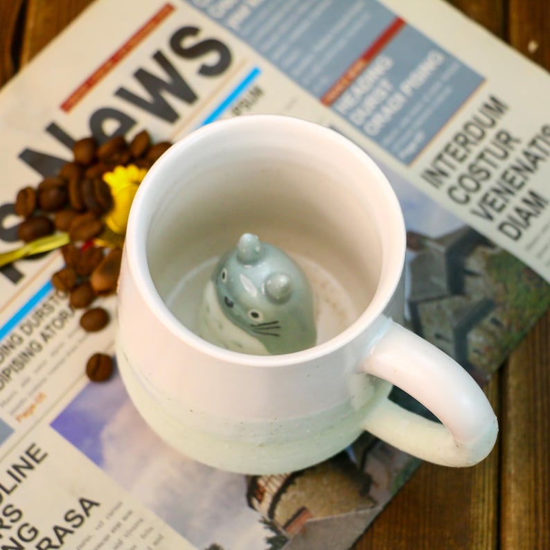 May include: A white ceramic mug with a light blue ombre design. A small ceramic figurine of a gray cat is inside the mug.