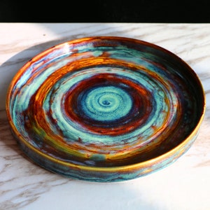 Può includere: Un piatto in ceramica fatto a mano con un design a spirale. Il piatto presenta una miscela di colori, tra cui verde acqua, marrone, arancione e blu. Il piatto è rotondo e ha un bordo leggermente rialzato. Il piatto ha un diametro di circa 25 cm.
