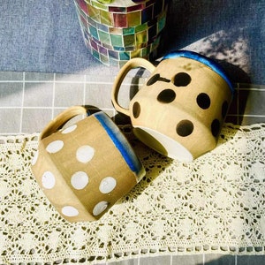 May include: Two ceramic mugs with polka dot patterns. One mug has white polka dots on a brown background, and the other has black polka dots on a brown background. Both mugs have a blue rim.