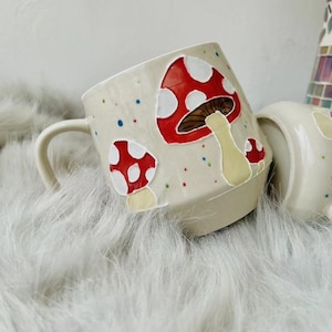 May include: A white ceramic mug with a handle, decorated with red and white mushrooms with brown stems. The mug has a speckled glaze with colourful dots.