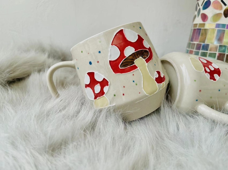 May include: A white ceramic mug with a handle, decorated with red and white mushrooms with brown stems. The mug has a speckled glaze with colourful dots.