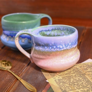 Puede incluir: Dos tazas de cerámica con un esmalte colorido. Las tazas son azules, verdes y rosas con un acabado moteado. Las tazas están sobre una superficie de madera.