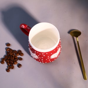 May include: Red ceramic mug with white polka dots and white heart designs. The mug is empty and sitting on a gray surface with a small pile of coffee beans next to it. A gold teaspoon is lying on the surface to the right of the mug.