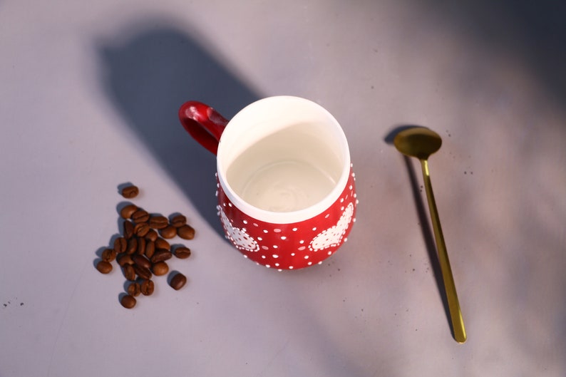 May include: Red ceramic mug with white polka dots and white heart designs. The mug is empty and sitting on a gray surface with a small pile of coffee beans next to it. A gold teaspoon is lying on the surface to the right of the mug.