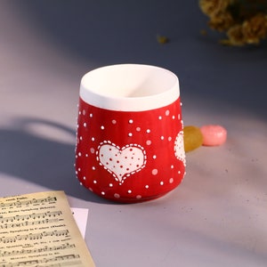 May include: A red ceramic mug with white polka dots and a white heart with white polka dots. The heart is in the center of the mug.