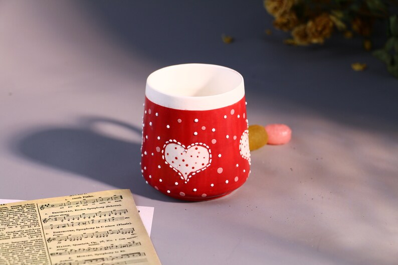 May include: A red ceramic mug with white polka dots and a white heart with white polka dots. The heart is in the center of the mug.