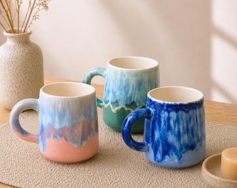 Op de afbeelding: Drie keramische mokken met een getextureerd, gedruppeld glazuureffect. De mokken zijn in tinten blauw, groen en roze, met een crèmekleurig interieur. Elke mok heeft een afgeronde handgreep. De mokken zijn gerangschikt op een neutraal gekleurde placemat.
