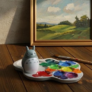 Puede incluir: Una paleta de cerámica con una variedad de colores de pintura, una figura de Totoro gris y blanco y un pincel. Una pintura al óleo de un paisaje está en el fondo. La paleta está sobre una superficie de madera.