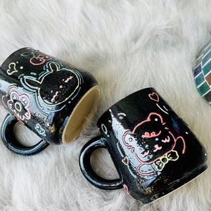 Peut inclure: Deux tasses en céramique noire avec des illustrations d'animaux de bande dessinée colorés. Une tasse représente un lapin avec des cerises et des fleurs, l'autre un ours avec des cœurs et des étoiles.