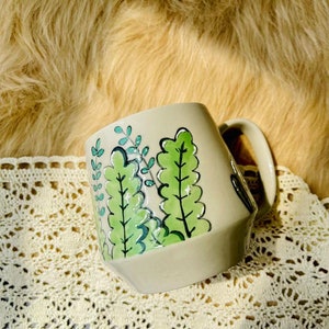 May include: A ceramic mug with a light gray background and a green leafy design. The mug has a handle and a rounded shape.