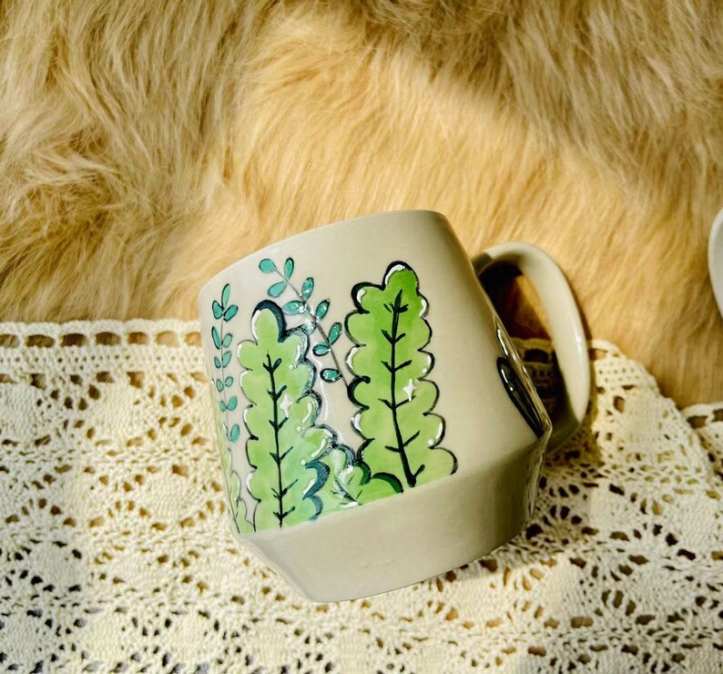 May include: A ceramic mug with a light gray background and a green leafy design. The mug has a handle and a rounded shape.