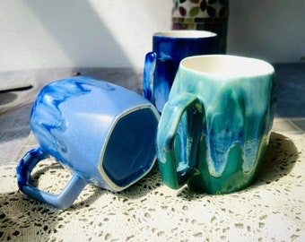 Op de afbeelding: Drie keramische mokken met een blauwe en groene glazuur. De mokken hebben een unieke, onregelmatige vorm en zijn perfect voor een warme drank.