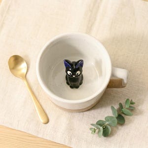 May include: A white ceramic mug with a black cat figurine inside. The mug has a tan base and handle. A gold spoon and sprig of green leaves are next to the mug on a beige surface.