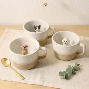 May include: Three white ceramic mugs with tan bases and handles, each containing a small animal figurine. One mug has a dog, another a cat, and the third a poodle. A gold spoon is placed on the table beside the mugs.