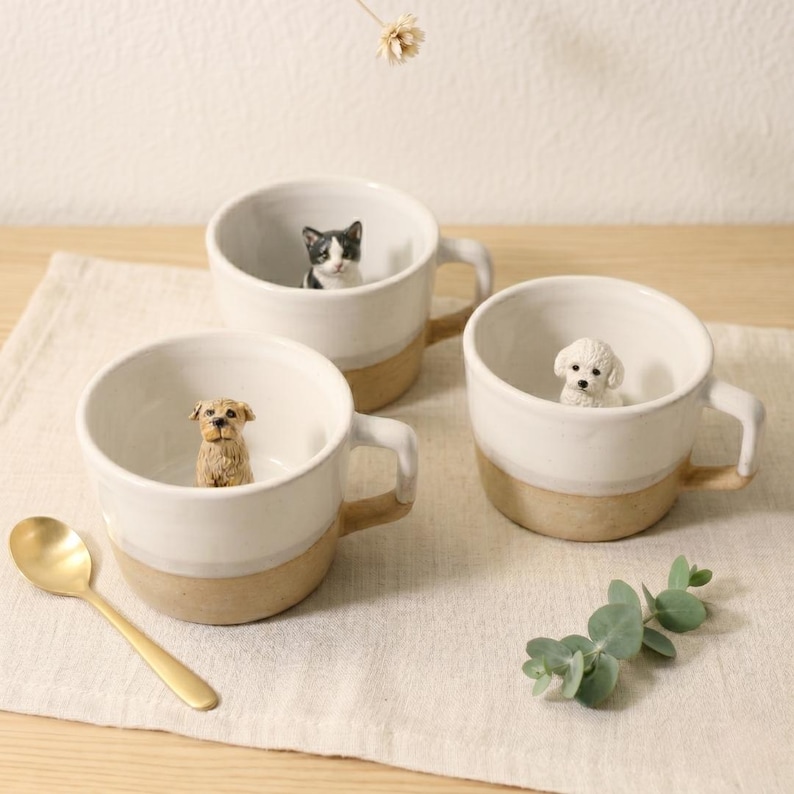 May include: Three white ceramic mugs with tan bases and handles, each containing a small animal figurine. One mug has a dog, another a cat, and the third a poodle. A gold spoon is placed on the table beside the mugs.