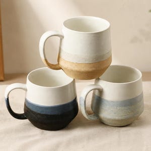 May include: Three ceramic mugs with a minimalist design. Each mug features a white upper section, a colored band in the middle, and a neutral-toned base. The mugs have large handles and a smooth, matte finish, perfect for coffee or tea.