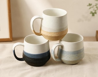 Puede incluir: Tres tazas de cerámica con un diseño minimalista. Cada taza presenta una sección superior blanca, una banda de color en el medio y una base de tonos neutros. Las tazas tienen asas grandes y un acabado liso y mate, para café o té.