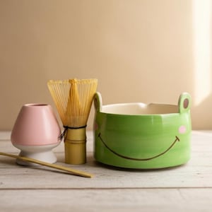 Könnte beinhalten: Ein Matcha-Teeset mit einer rosa Keramik-Teeschale, einem Bambusbesen, einer grünen froschförmigen Schale mit einem lächelnden Gesicht und einem Bambuslöffel. Das Set ist auf einer hellen Holzoberfläche angeordnet.
