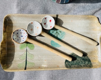 May include: Three ceramic spoons with different patterns on a rectangular ceramic tray with a green and blue floral design. The spoons are white with red, blue, and white polka dots.