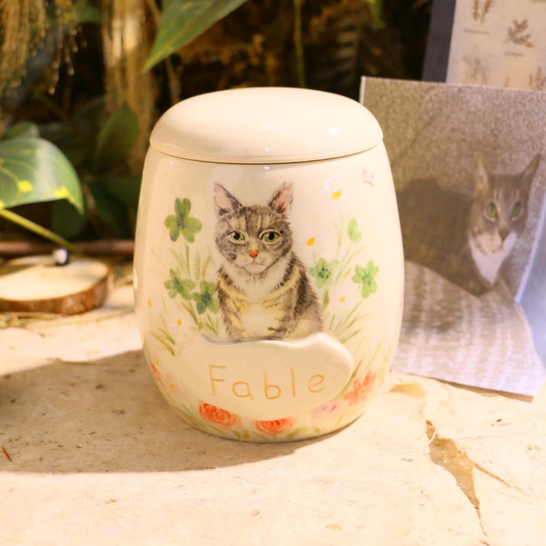 Puede incluir: Un tarro de cer&aacute;mica color crema con tapa a juego, que presenta una ilustraci&oacute;n pintada a mano de un gato atigrado rodeado de tr&eacute;boles verdes y peque&ntilde;as flores naranjas. La palabra Fable est&aacute; impresa en color marr&oacute;n claro sobre un gr&aacute;fico de nube.