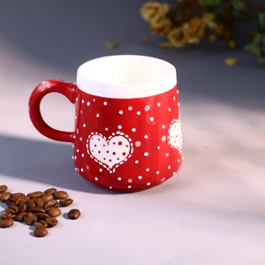 May include: Red ceramic mug with white polka dots and a white heart design. The heart has white polka dots inside.