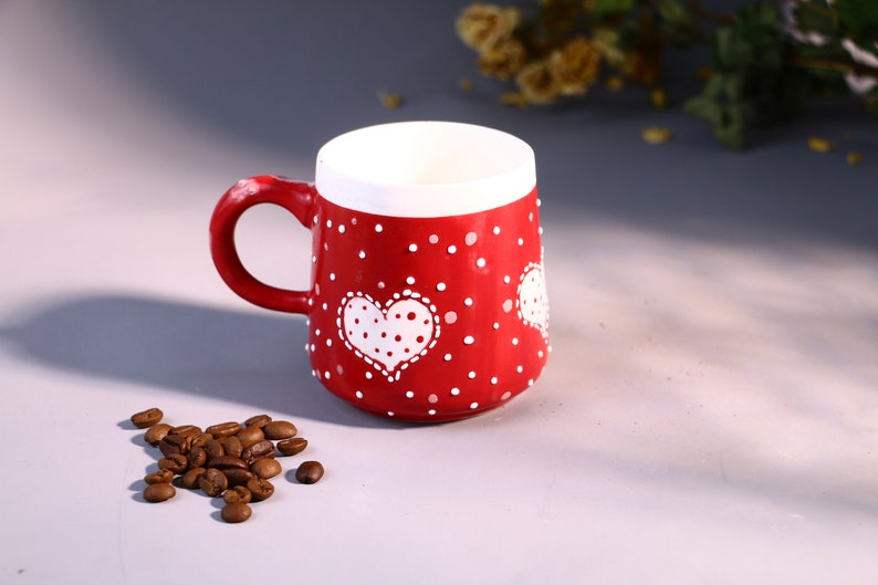 May include: Red ceramic mug with white polka dots and a white heart design. The heart has white polka dots inside.